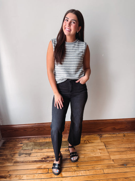 A woman modeling a sleeveless gray and white striped crop top with black cropped jeans and black slide sandals, standing on a wooden floor against a plain off-white wall.