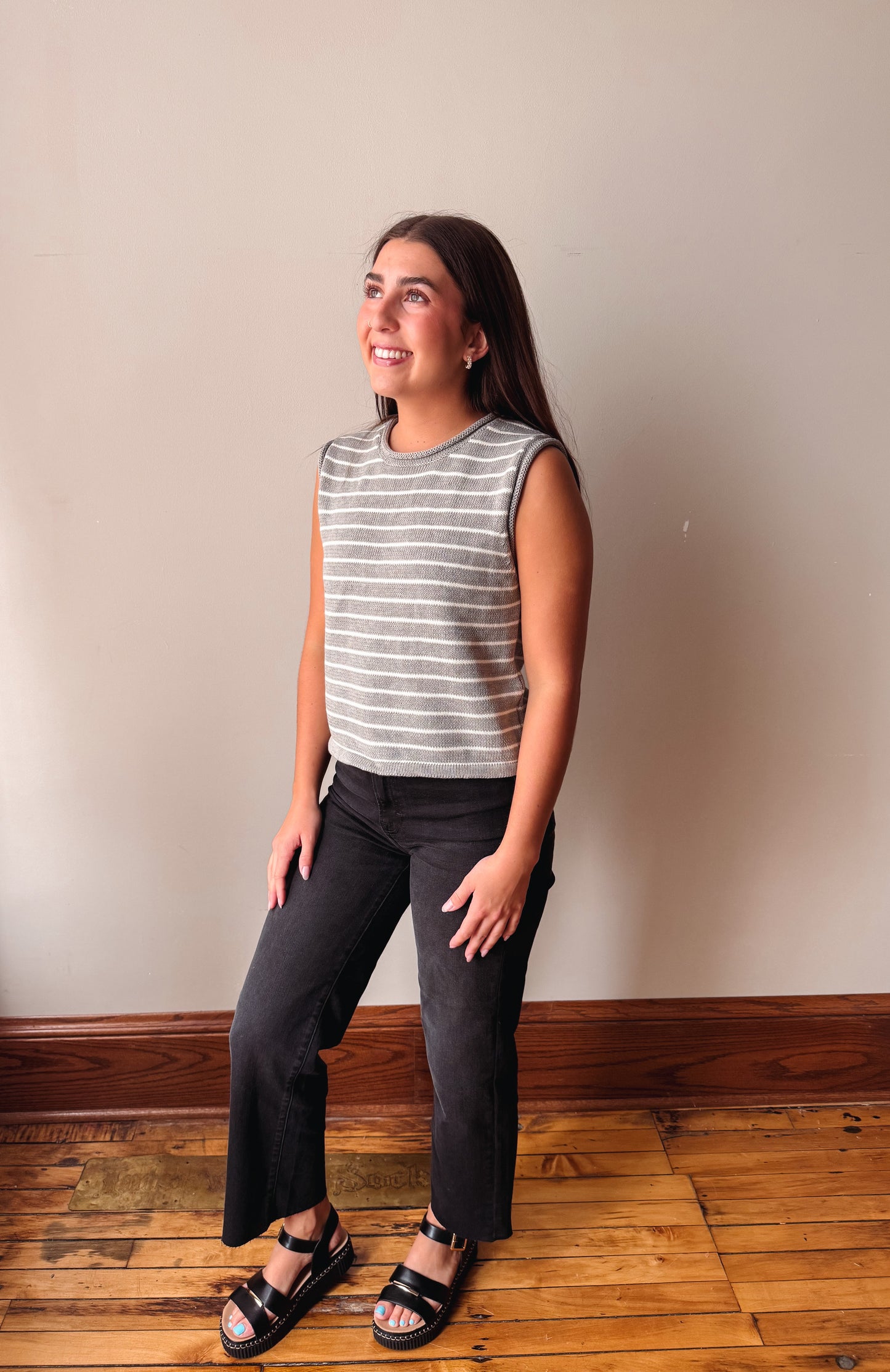 A woman modeling a sleeveless gray and white striped knit top with dark charcoal pants and black sandals, standing on a wooden floor against a plain beige wall.