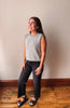 A woman modeling a sleeveless gray and white striped knit top with dark charcoal pants and black sandals, standing on a wooden floor against a plain beige wall.