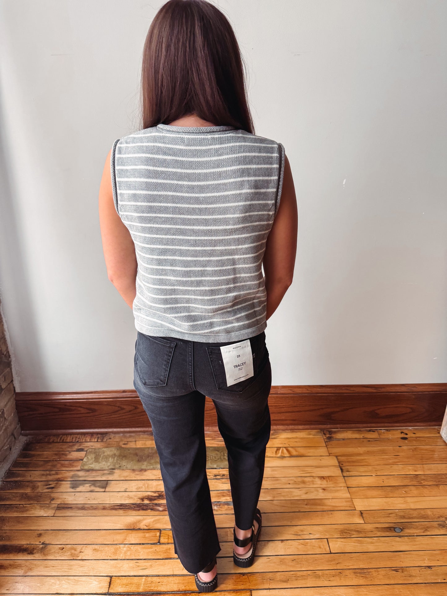 A woman viewed from behind wearing a gray and white striped sleeveless top and dark charcoal gray jeans, standing on a wooden floor against a white wall.