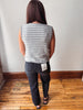 A woman viewed from behind wearing a gray and white striped sleeveless top and dark charcoal gray jeans, standing on a wooden floor against a white wall.