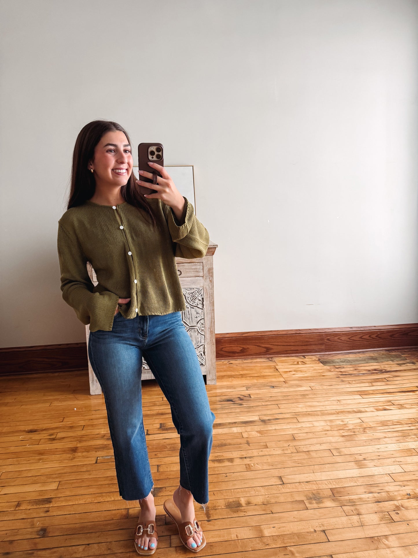 A woman takes a mirror selfie wearing an olive green button-up knit cardigan, blue jeans, and pink mule sandals in a room with a wooden floor and white wall.