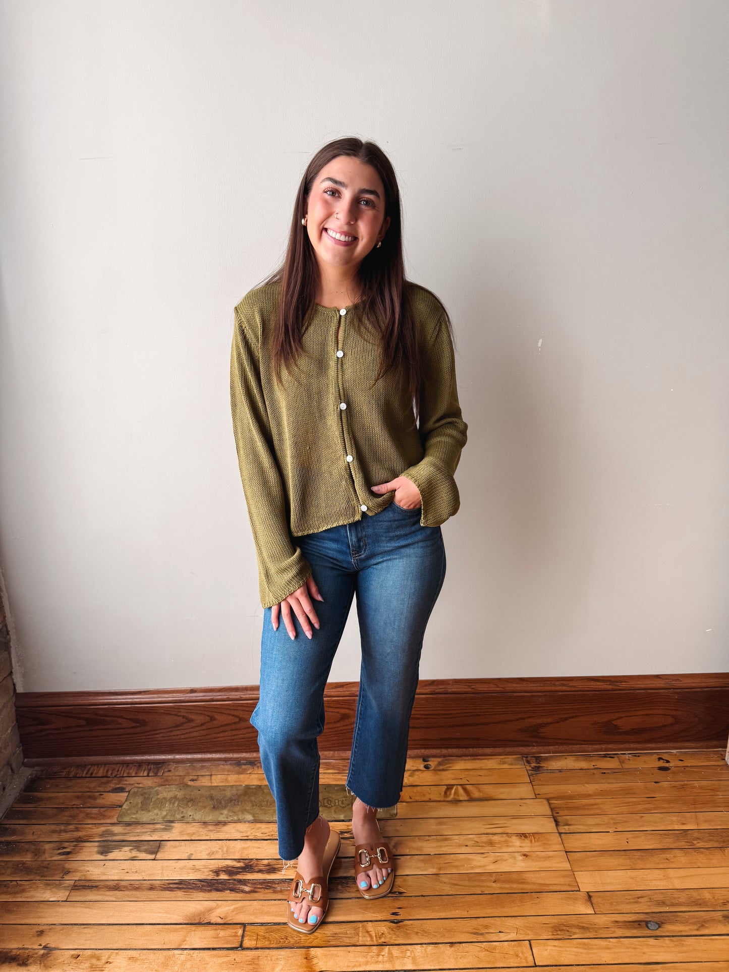 A woman stands in a room with a white wall and wooden floor, wearing an olive green cropped button-up cardigan, blue jeans, and brown flat sandals.