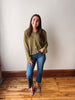 A woman stands in a room with a white wall and wooden floor, wearing an olive green cropped button-up cardigan, blue jeans, and brown flat sandals.