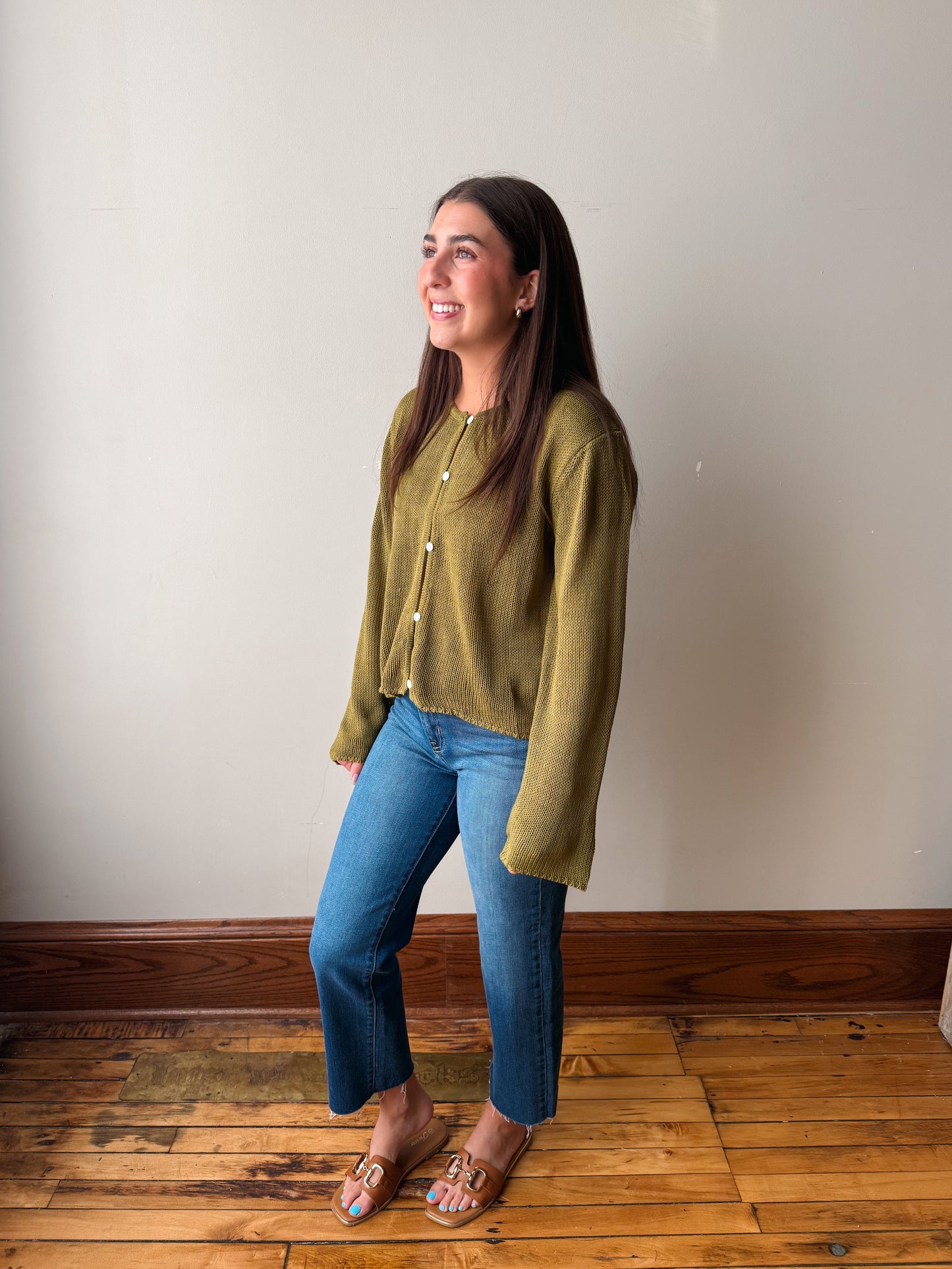 A woman wearing a textured olive green knit cardigan with gold buttons, blue jeans, and flat sandals, standing on a wooden floor against a plain off-white wall.