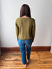 A woman viewed from behind wearing a textured olive green long-sleeve sweater and blue jeans, standing on a wooden floor against a plain white wall.