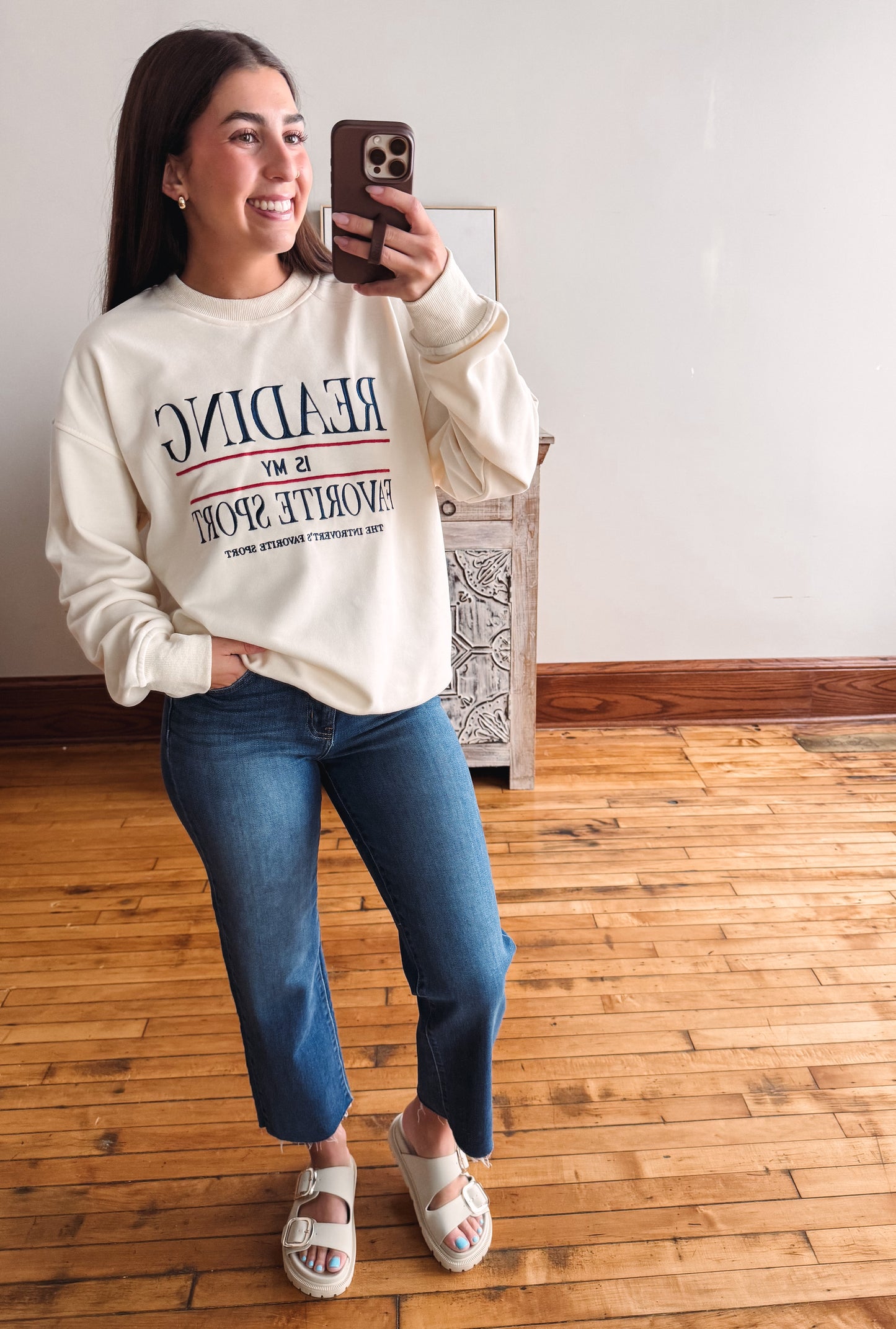 A woman taking a mirror selfie wearing a cream sweatshirt with navy and red text that reads 'READING IS MY FAVORITE SPORT', blue jeans, and white slide sandals in a room with wooden floors.