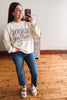 A woman taking a mirror selfie wearing a cream sweatshirt with navy and red text that reads 'READING IS MY FAVORITE SPORT', blue jeans, and white slide sandals in a room with wooden floors.