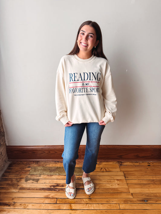 A woman stands indoors wearing a cream sweatshirt with blue and red text reading 'READING IS MY FAVORITE SPORT,' blue jeans, and white slide sandals. The photo is taken against a plain light wall with a wooden floor.