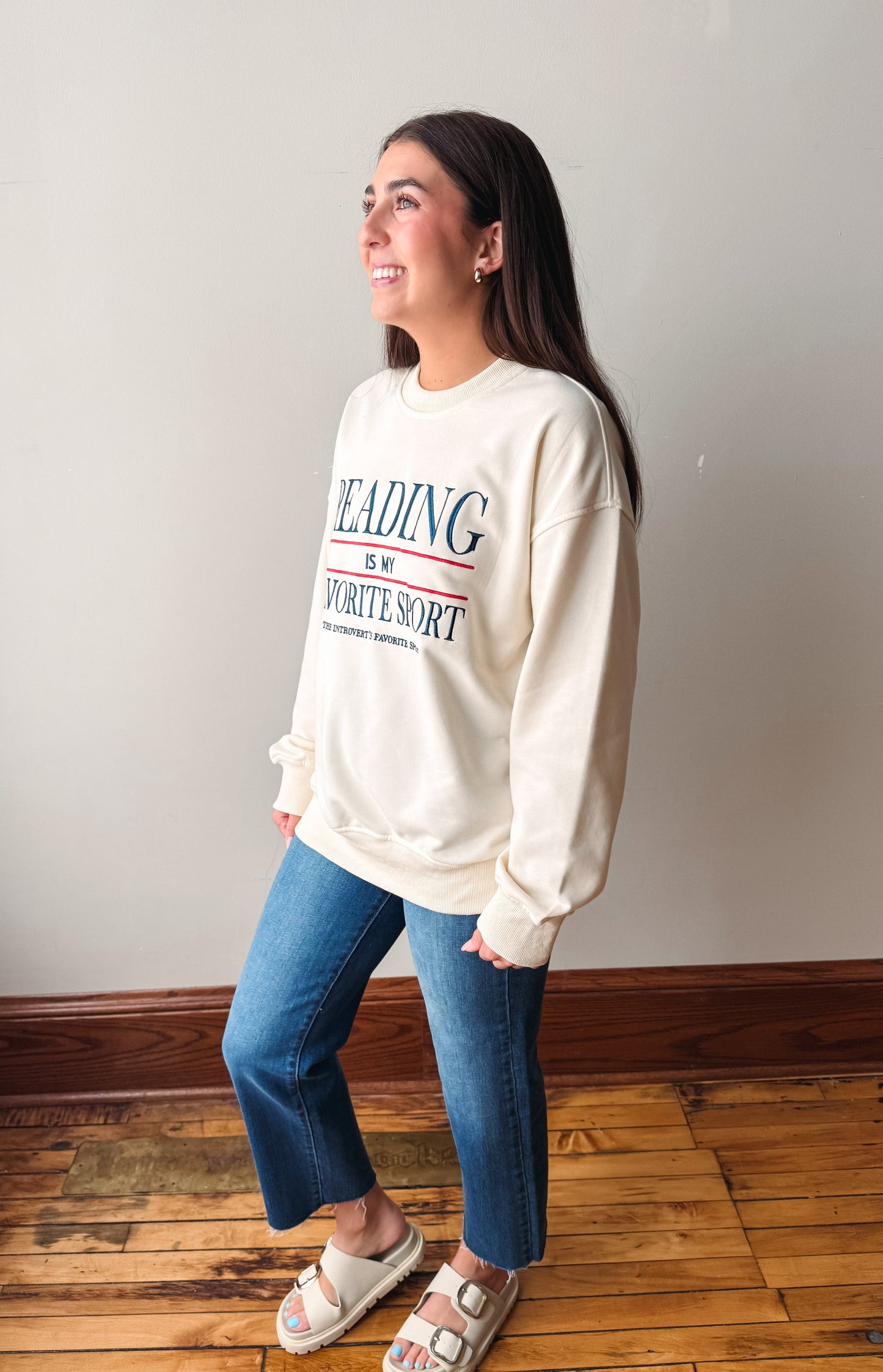 A woman wearing a cream-colored crewneck sweatshirt with blue and red text that reads 'READING IS MY WRITE ART', paired with blue jeans and white slide sandals, standing on a wooden floor against a plain wall.