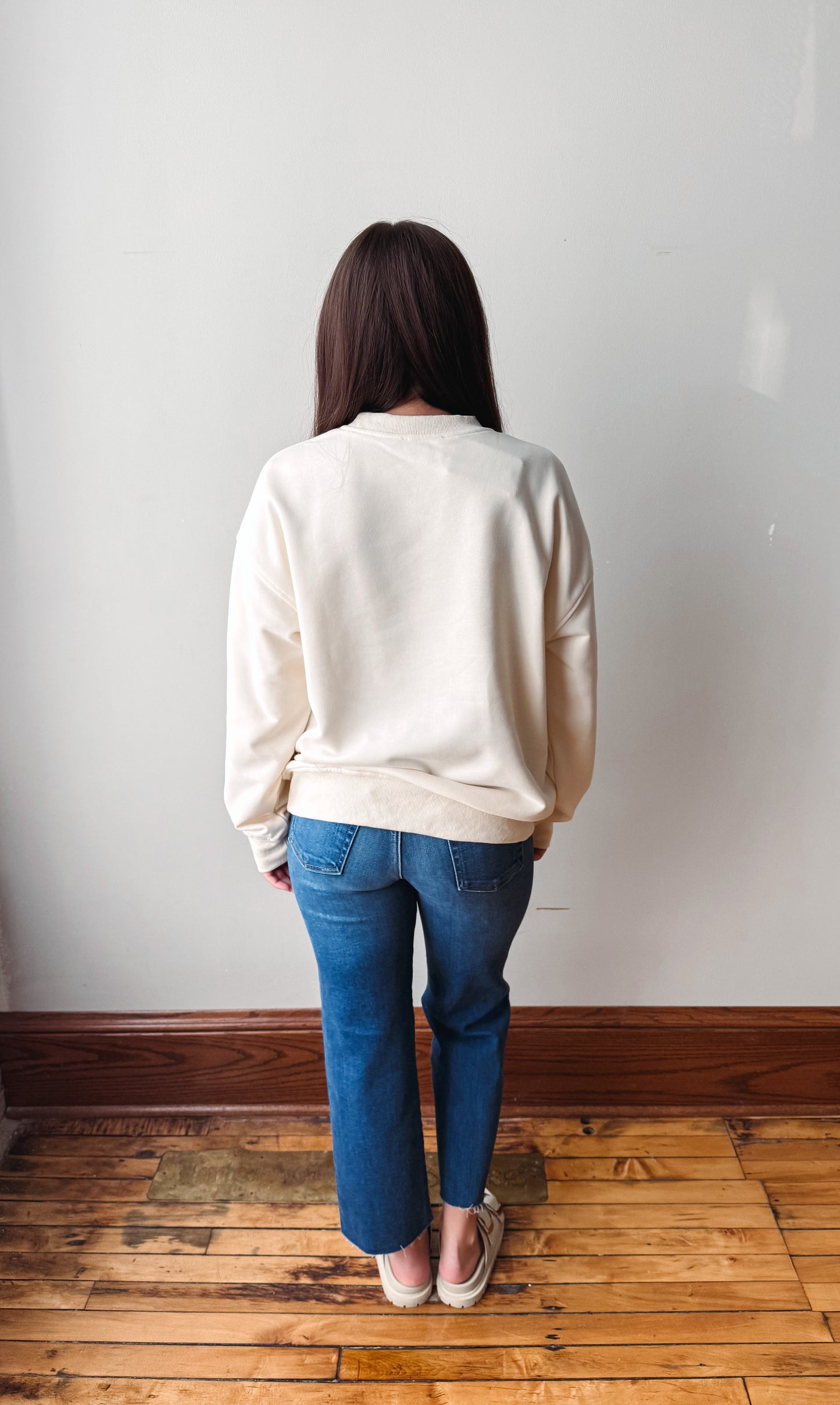 A woman viewed from behind wearing a cream-colored crewneck sweatshirt and blue jeans, standing on a wooden floor against a plain white wall.