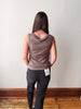 A woman viewed from behind wearing a sleeveless, cowl-neck taupe top and dark gray jeans, standing on a wooden floor against a plain off-white wall.