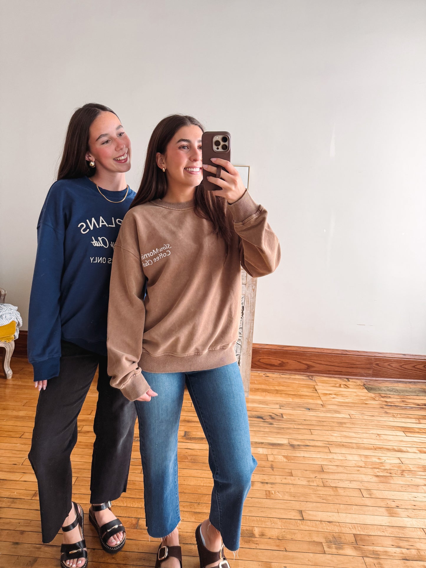Two women taking a mirror selfie indoors. The woman in front wears a taupe crewneck sweatshirt and blue jeans, while the woman behind wears a navy sweatshirt with white text and dark pants.
