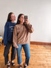 Two women taking a mirror selfie indoors. The woman in front wears a taupe crewneck sweatshirt and blue jeans, while the woman behind wears a navy sweatshirt with white text and dark pants.