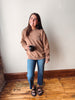 A woman stands indoors against a plain off-white wall on a wood floor, wearing a tan crewneck sweatshirt with a small embroidered logo on the left chest, blue jeans, and black slide sandals while holding a black mug.