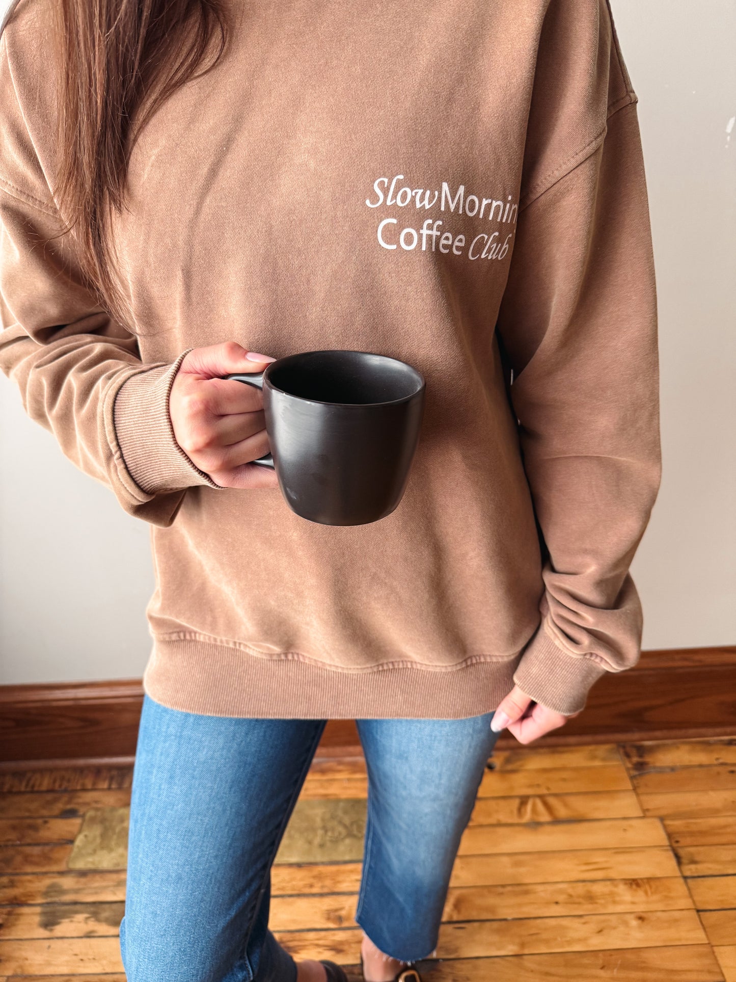 A person wearing an oversized tan sweatshirt with the text 'SlowMornin Coffee Club' printed in white on the chest, holding a black ceramic mug. The person is also wearing blue jeans, standing on a wooden floor against a plain wall.