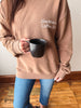 A person wearing an oversized tan sweatshirt with the text 'SlowMornin Coffee Club' printed in white on the chest, holding a black ceramic mug. The person is also wearing blue jeans, standing on a wooden floor against a plain wall.