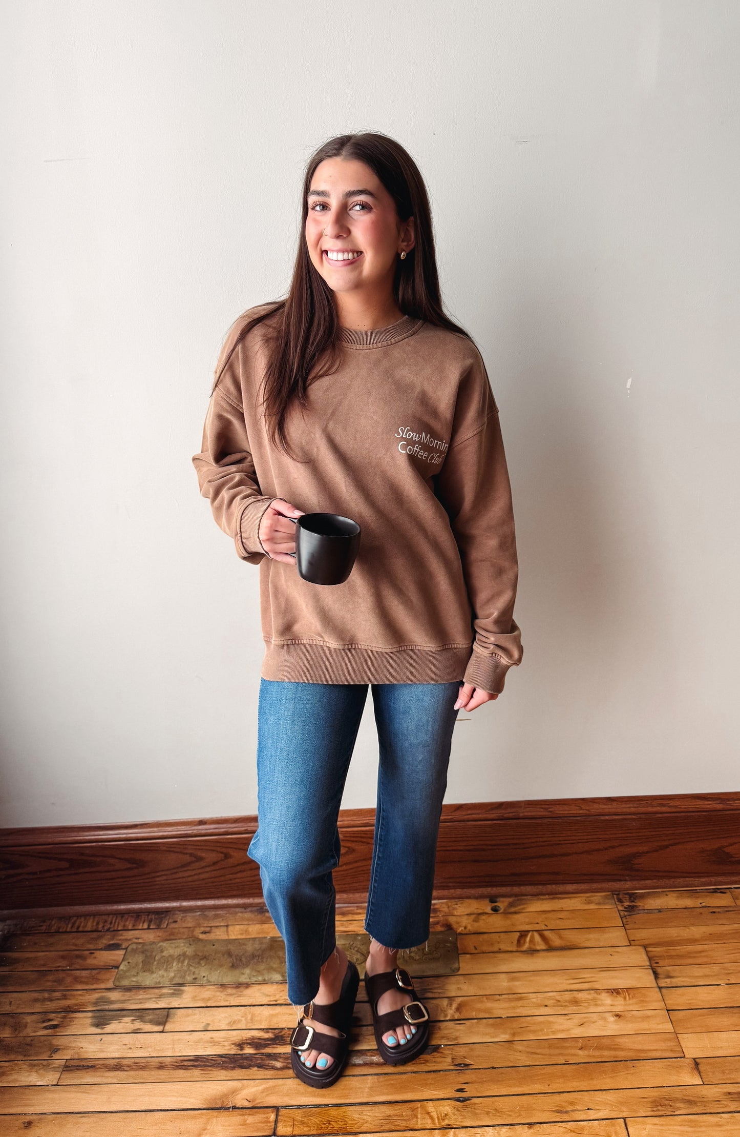 A woman stands indoors wearing a brown crewneck sweatshirt with a small embroidered logo on the chest, blue jeans, and brown sandals, while holding a black mug against a plain off-white wall and wooden floor.