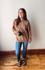 A woman stands indoors wearing a brown crewneck sweatshirt with a small embroidered logo on the chest, blue jeans, and brown sandals, while holding a black mug against a plain off-white wall and wooden floor.