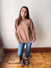 A woman stands indoors wearing a taupe crewneck sweatshirt with 'Slow morning coffee club' printed on the chest, blue jeans, and brown sandals, posing against a white wall and wooden floor.