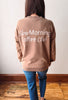 A woman viewed from behind wearing a brown sweatshirt with 'Slow Morning Coffee Club' printed in white script across the back, paired with blue jeans, standing on a wooden floor against a plain wall.