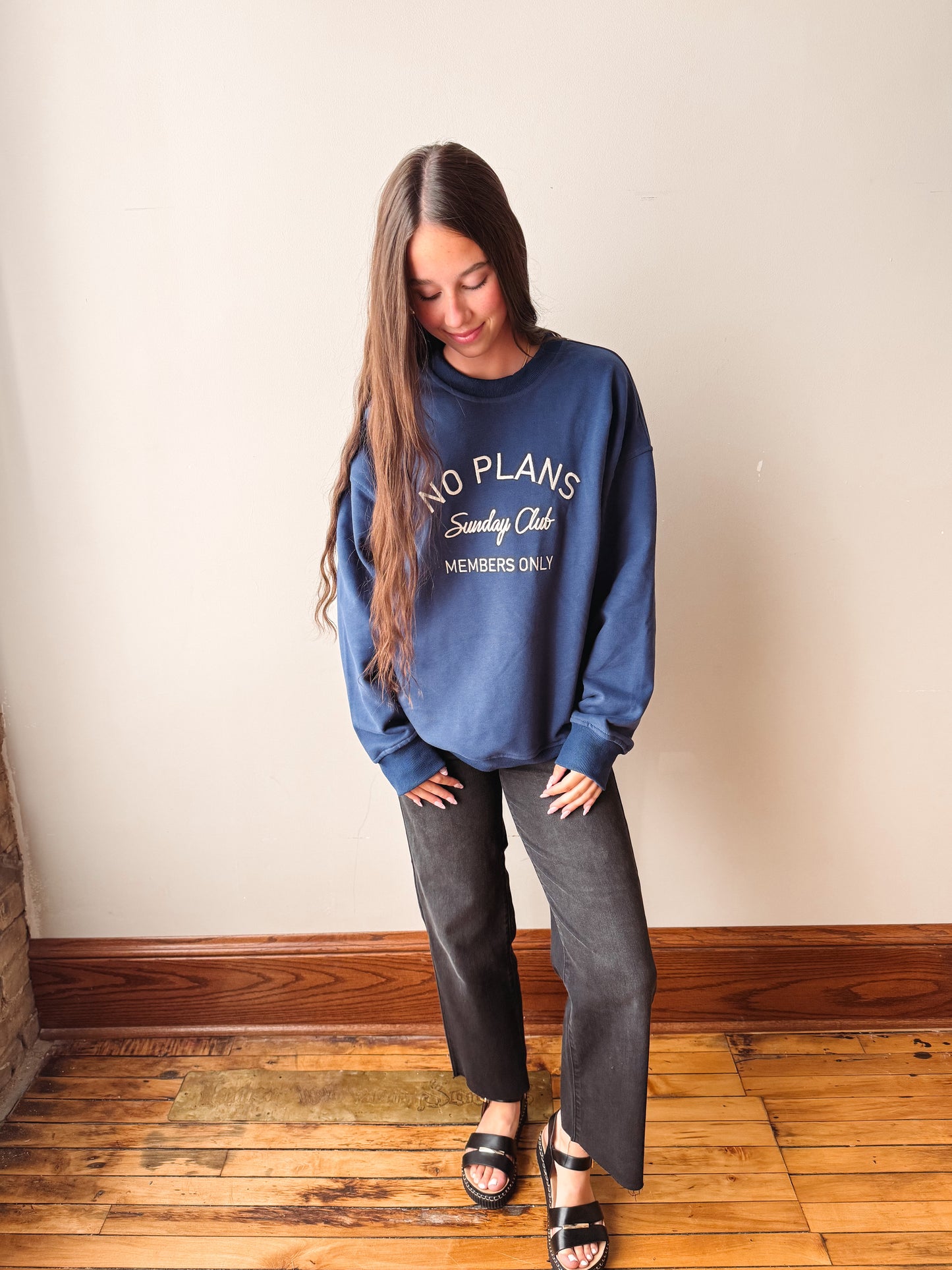 A woman wearing a blue crewneck sweatshirt with white text that reads 'NO PLANS Sunday Club MEMBERS ONLY', paired with dark gray wide-leg pants and black sandals, standing on a wooden floor against a beige wall.