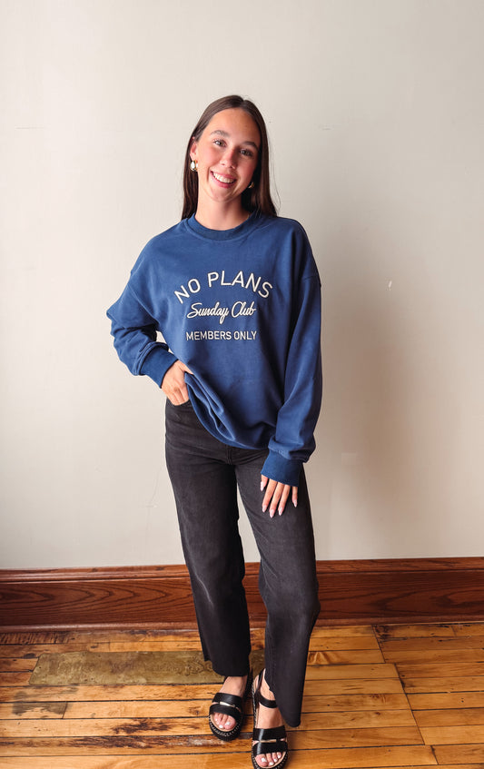 A woman stands indoors wearing a navy blue sweatshirt with white text reading 'NO PLANS Sunday Club MEMBERS ONLY,' paired with dark pants and black sandals, against a plain wall and wood floor.
