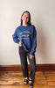 A woman stands indoors wearing a navy blue sweatshirt with white text reading 'NO PLANS Sunday Club MEMBERS ONLY,' paired with dark pants and black sandals, against a plain wall and wood floor.