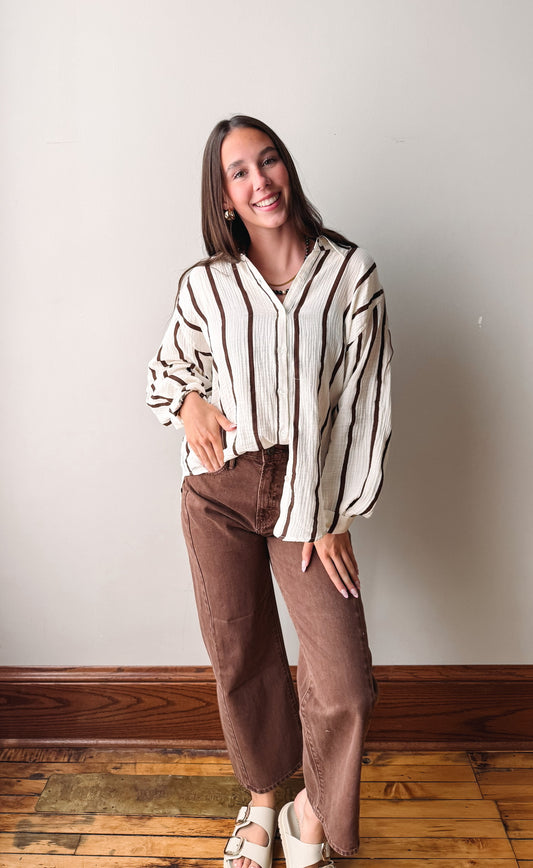 A woman modeling a cream and brown vertically striped long-sleeve button up with brown pants, standing against a plain light gray wall on a wooden floor.