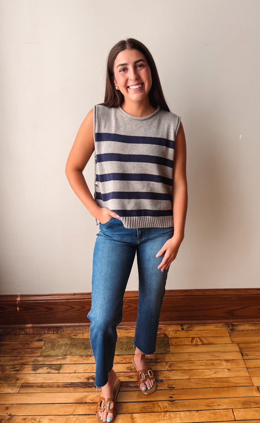 A woman modeling a sleeveless knit gray sweater vest with horizontal navy stripes, paired with blue jeans and flat sandals, standing on a wooden floor against a plain beige wall.