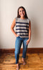 A woman modeling a sleeveless knit gray sweater vest with horizontal navy stripes, paired with blue jeans and flat sandals, standing on a wooden floor against a plain beige wall.