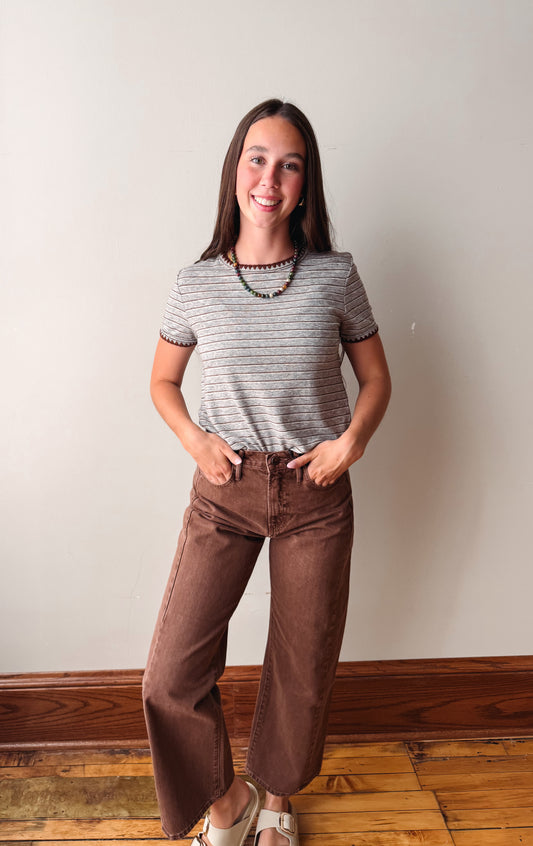 A young woman stands indoors against a plain off-white wall, wearing a short-sleeve gray and brown striped t-shirt, brown wide-leg pants, and white slide sandals.