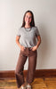 A young woman stands indoors against a plain off-white wall, wearing a short-sleeve gray and brown striped t-shirt, brown wide-leg pants, and white slide sandals.