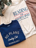 Three crewneck sweatshirts arranged in a flat lay on a light wood surface: a navy blue sweatshirt in front reading 'NO PLANS Sunday Club MEMBERS ONLY' in white, a cream sweatshirt behind it reading 'READING IS MY FAVORITE SPORT' in navy and red, and part of a brown sweatshirt at the top right with partial white text.