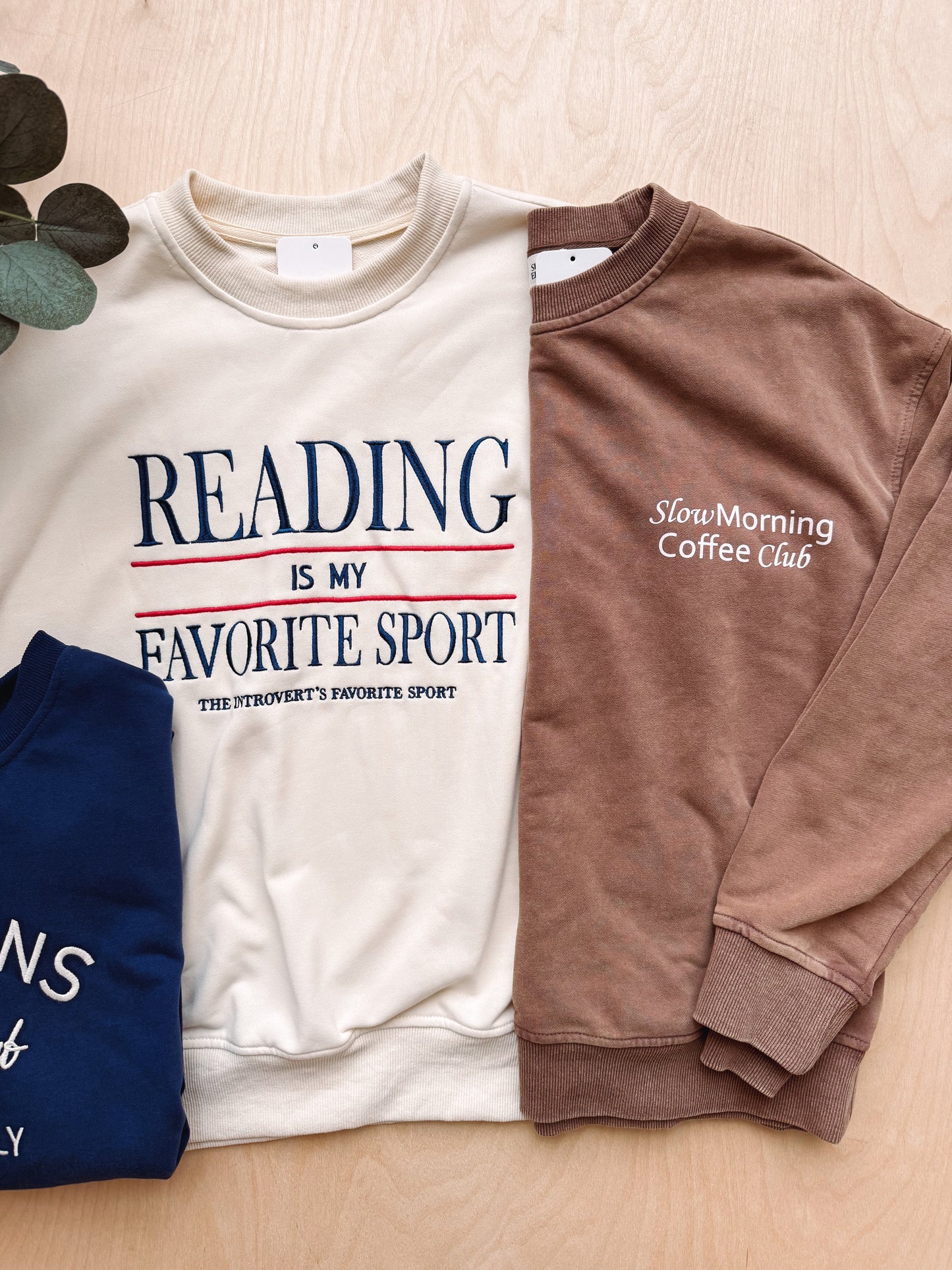Three crewneck sweatshirts laid flat on a light wood surface: an off-white sweatshirt with navy and red text reading 'READING IS MY FAVORITE SPORT,' a brown sweatshirt with white text reading 'SlowMorning Coffee Club,' and a navy sweatshirt partially visible on the left. A leafy stem decorates the top left corner.