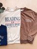 Three crewneck sweatshirts laid flat on a light wood surface: an off-white sweatshirt with navy and red text reading 'READING IS MY FAVORITE SPORT,' a brown sweatshirt with white text reading 'SlowMorning Coffee Club,' and a navy sweatshirt partially visible on the left. A leafy stem decorates the top left corner.