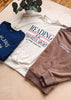 Three crewneck sweatshirts in navy blue, cream, and taupe brown are arranged in a flat lay on a light wood background. The cream sweatshirt in the center features a graphic print that reads 'READING IS MY FAVORITE SPORT'.
