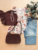 A styled flat-lay outfit on a light wood background featuring a dark brown ruffled top, a cream floral blouse, light blue rolled-calf jeans, a dark brown woven crossbody bag, a white beaded necklace, and a potted eucalyptus plant.