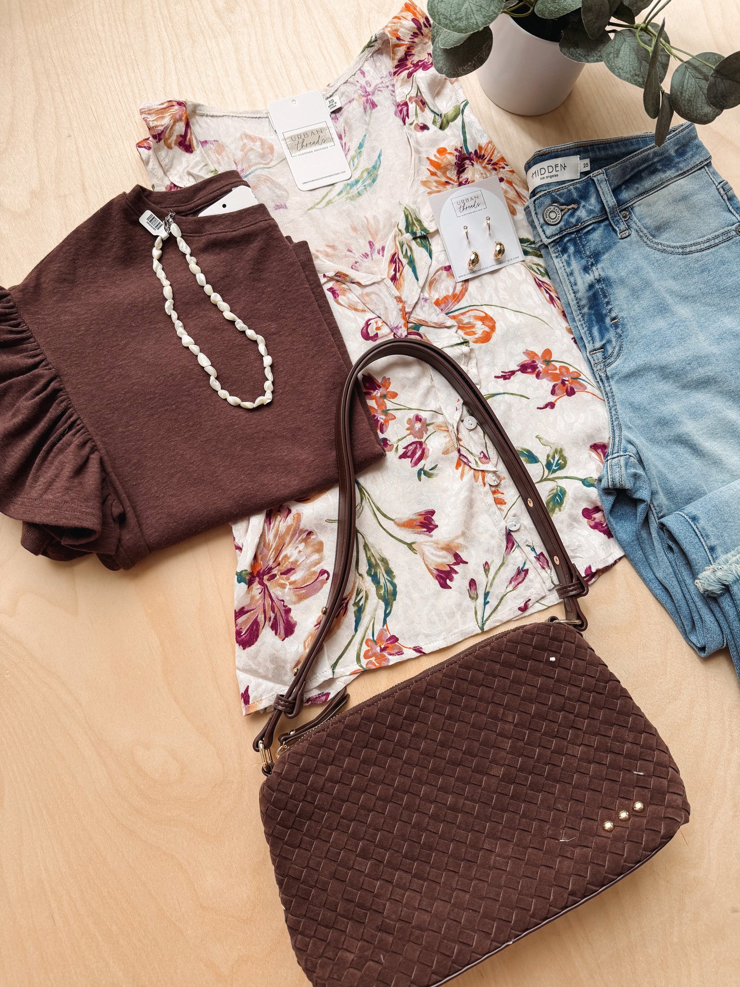A styled flat-lay of a clothing and accessory set on a light wood surface, featuring a dark brown ruffled top, a cream floral long-sleeve top, light blue denim shorts, a dark brown woven crossbody bag, and a white beaded necklace.