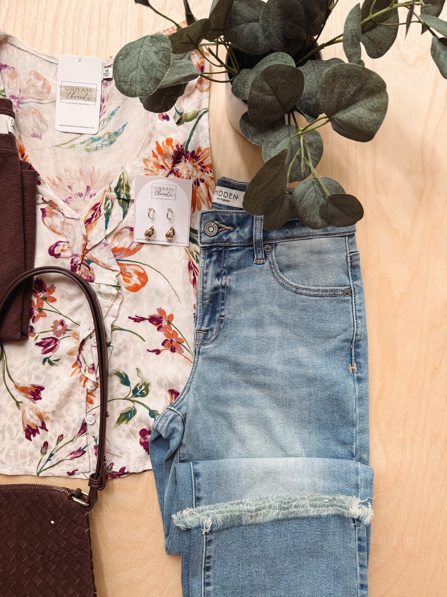 A styled flat-lay featuring a pair of light-wash blue denim jeans with rolled cuffs and raw frayed hems, laid next to a cream floral short-sleeve top and a dark brown woven handbag on a light wood background with eucalyptus leaves.