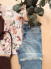 A styled flat-lay featuring a pair of light-wash blue denim jeans with rolled cuffs and raw frayed hems, laid next to a cream floral short-sleeve top and a dark brown woven handbag on a light wood background with eucalyptus leaves.