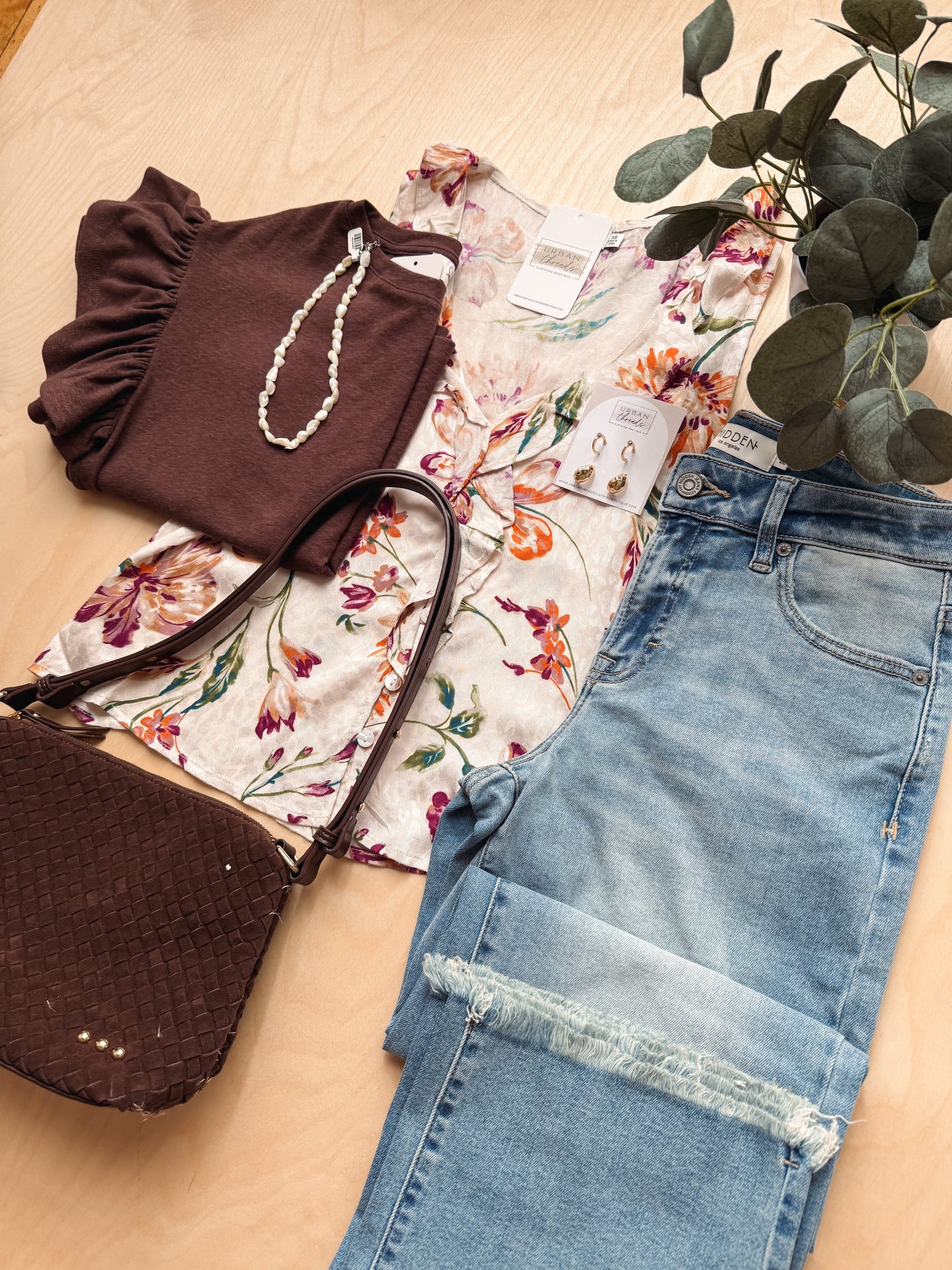 A styled flat-lay of clothing and accessories on a light wood surface: a brown ruffled top, a cream floral blouse, light blue distressed jeans, a dark brown woven handbag, a pearl necklace, and a green leafy plant.