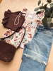 A styled flat-lay of clothing and accessories on a light wood surface: a brown ruffled top, a cream floral blouse, light blue distressed jeans, a dark brown woven handbag, a pearl necklace, and a green leafy plant.