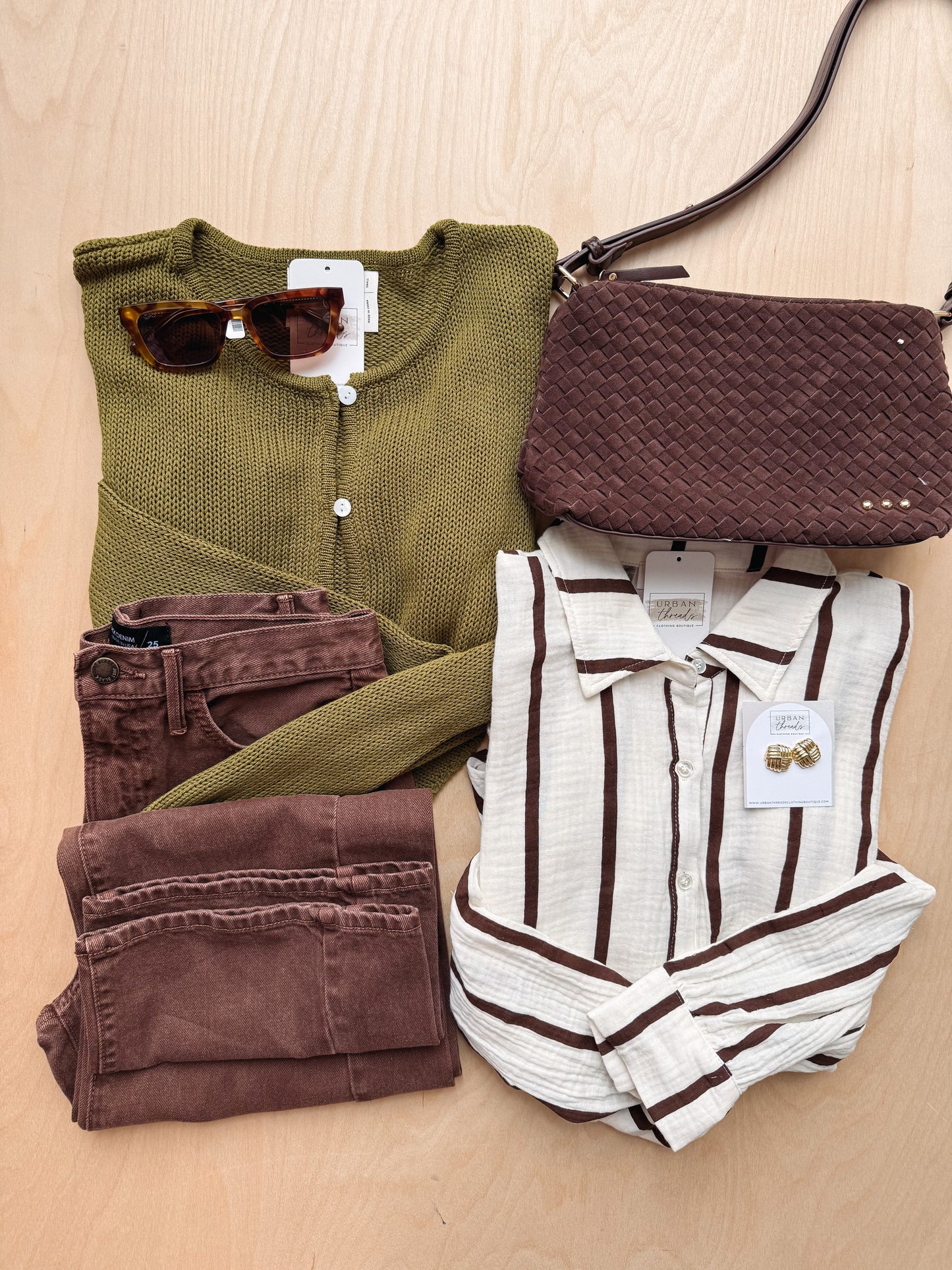 A styled flat-lay of multiple fashion items on a light wood background: an olive green knit top, brown pants, a brown woven crossbody bag, a cream and brown striped button-up shirt, sunglasses, and a pair of brown earrings.