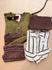 A styled flat-lay of multiple fashion items on a light wood background: an olive green knit top, brown pants, a brown woven crossbody bag, a cream and brown striped button-up shirt, sunglasses, and a pair of brown earrings.