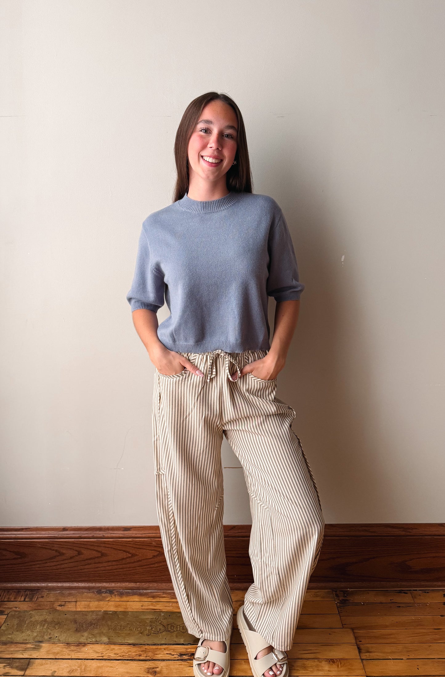 A woman stands against a plain beige wall on a wooden floor, wearing a light blue crewneck knit sweater and beige pinstriped wide-leg pants.