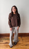 A woman stands in a room with a light gray wall and wooden floor, wearing a brown knit crewneck sweater and beige-and-black gingham flared pants with ruffled hems.