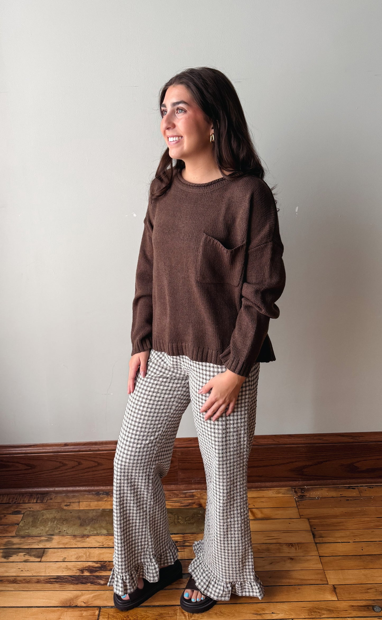 A woman modeling a brown long-sleeve knit top and black-and-white checkered flared pants, standing on a wooden floor against a plain light gray wall.