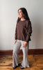 A woman modeling a brown long-sleeve knit top and black-and-white checkered flared pants, standing on a wooden floor against a plain light gray wall.