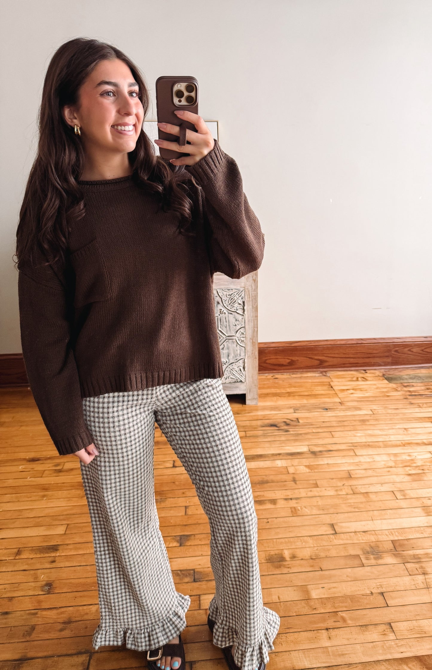 A woman takes a mirror selfie indoors wearing a dark brown knit sweater and black-and-white gingham flared pants with ruffled hems.